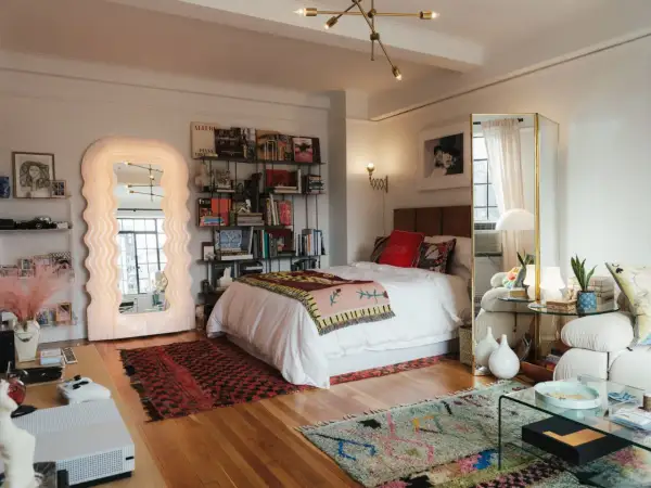 Un dormitorio con un diseño estudio moderno. Cama blanca, estanterías llenas de libros, un espejo ondulado y alfombras coloridas.