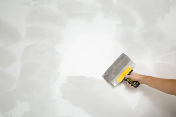 Mano con espátula amarilla y gris trabajando en una pared, alisando la textura para como quitar gotele.