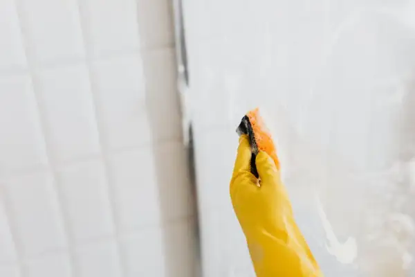 Mano con guante amarillo limpiando mampara de ducha con esponja.