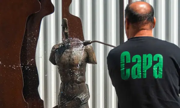 Hombre con camiseta negra con letras verdes "Capa" limpia una estatua de bronce con agua a presión.