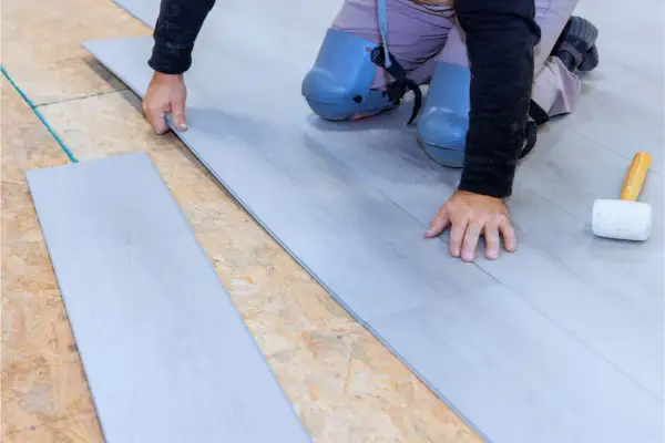 Man con rodilleras coloca cuidadosamente tablas de suelo laminado gris sobre una base de madera.