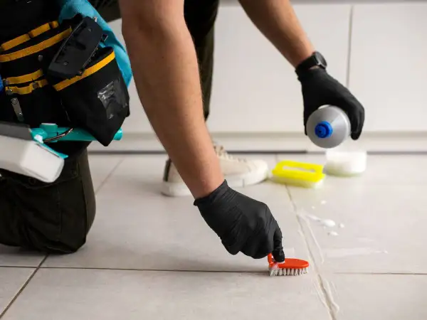 Persona con guantes negros frota las juntas de los azulejos con un cepillo naranja, aplicando un producto para blanquear juntas azulejos.
