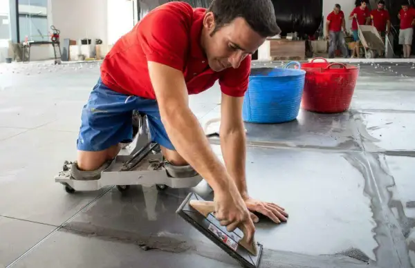 Un trabajador con rodilleras aplica cemento para saber como poner baldosas en el suelo.